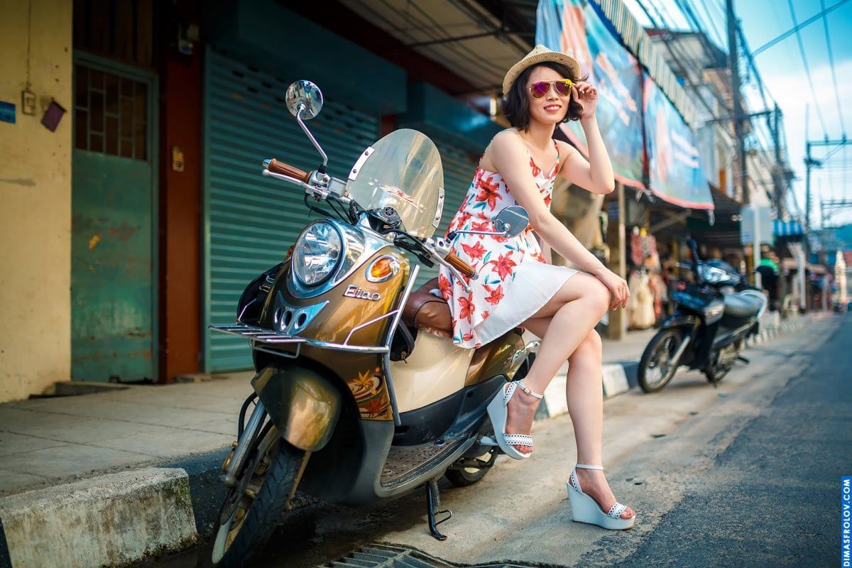 Joyful traveler in a summer dress posing on a scooter in a colorful street