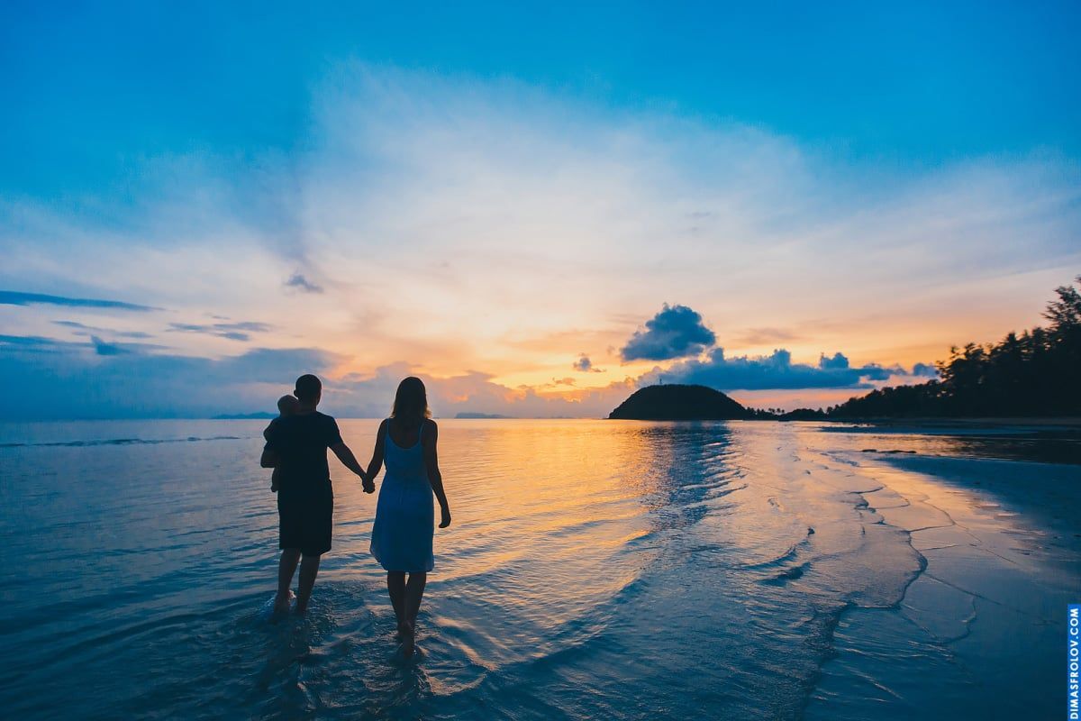 Sunset family photo shoot — parents walking in shallow sea with baby in dad’s arms