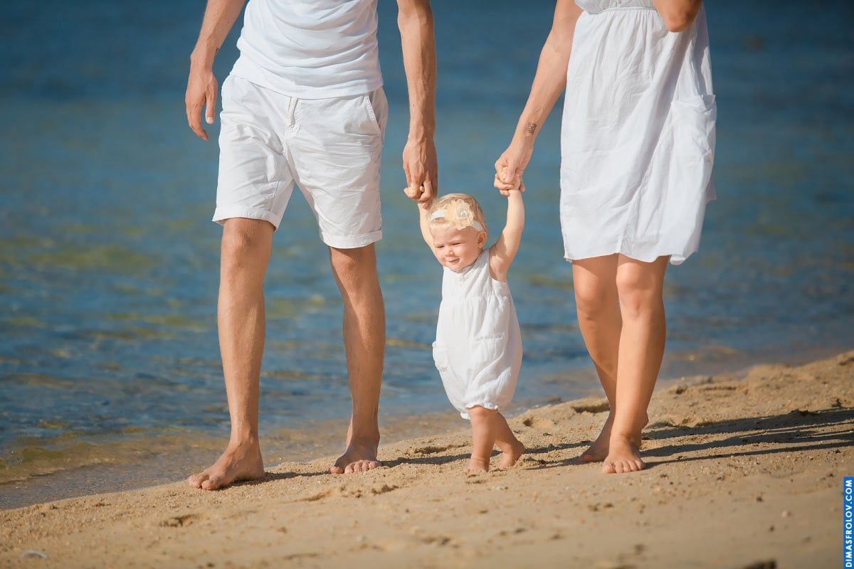 Candid family photo — toddler holding parents’ hands walking by the shore