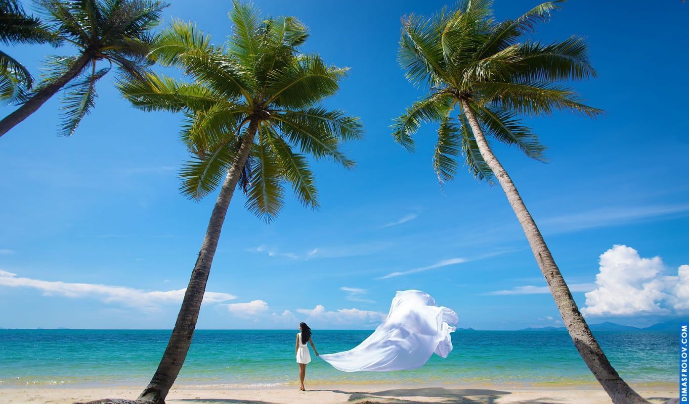 Woman in white with flowing fabric dancing in the ocean breeze between two palms