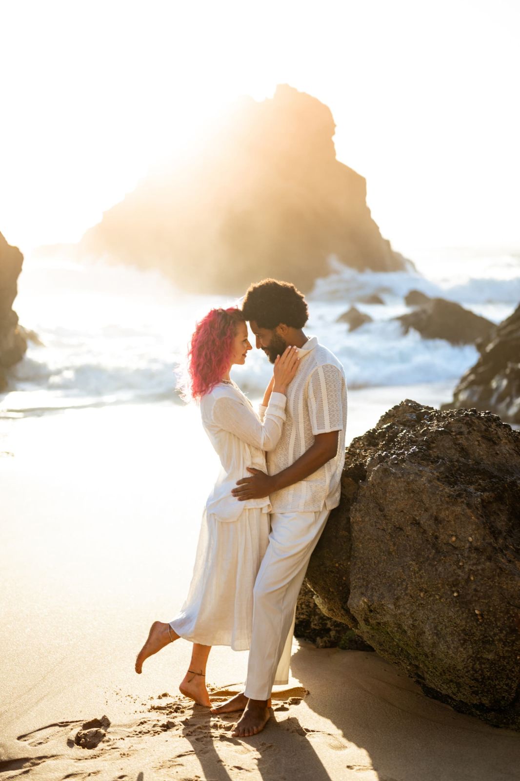Couple standing forehead to forehead against a rock on the sand at sunset
