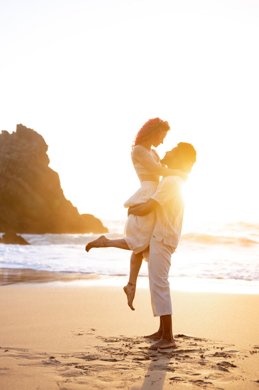 Man lifting his partner on the sand as the sun sets behind a sea cliff