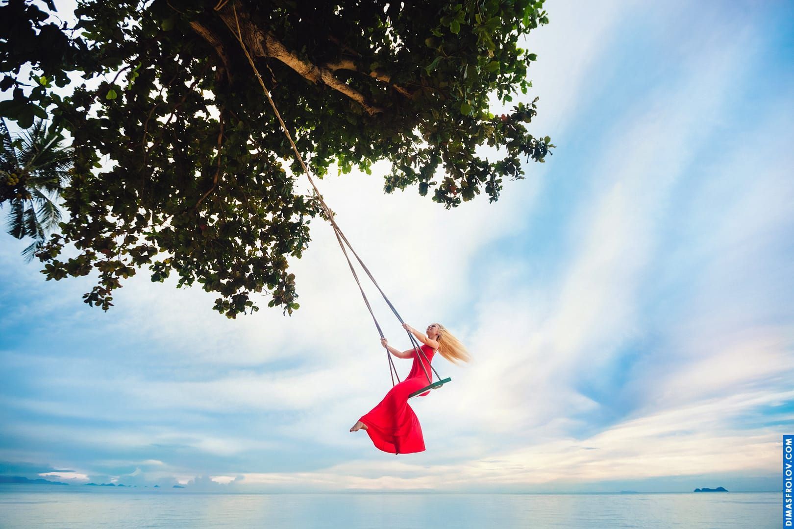 Magical swing photo of a girl in a red dress floating above the ocean under dreamy sky