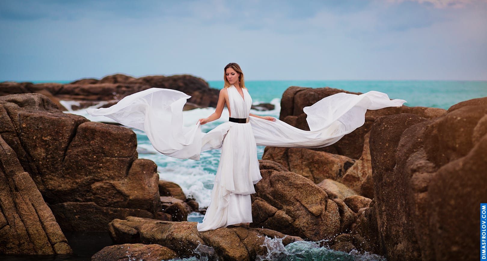 Dramatic seaside portrait of a woman in white dress on rocks with flowing fabric