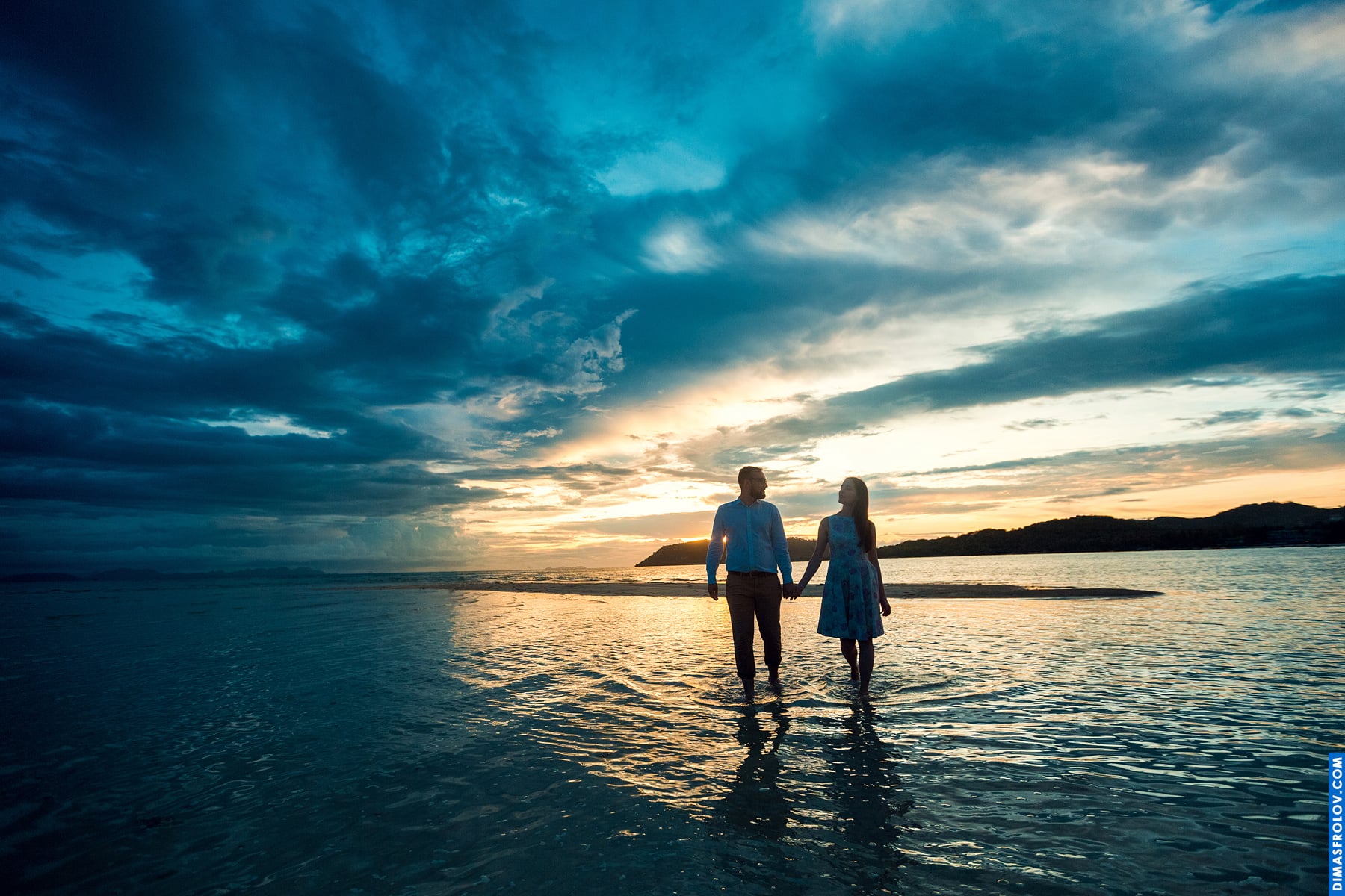 Couple walking through calm water under a dramatic sky, with mirrored clouds and wide-angle depth