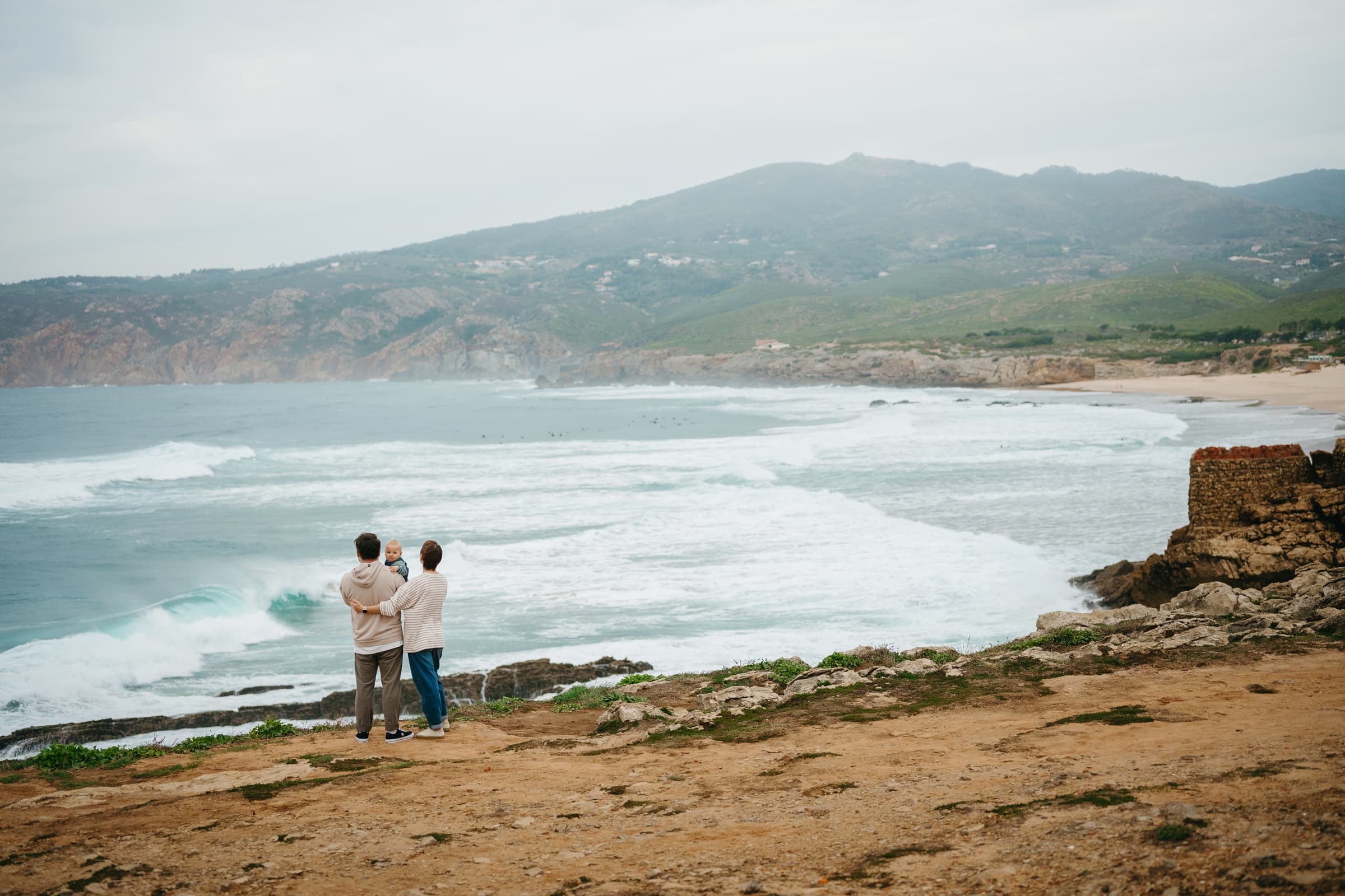 Parents holding a baby on the headland above Guincho beach with the Sintra hills behind, family photoshoot on the Atlantic coast