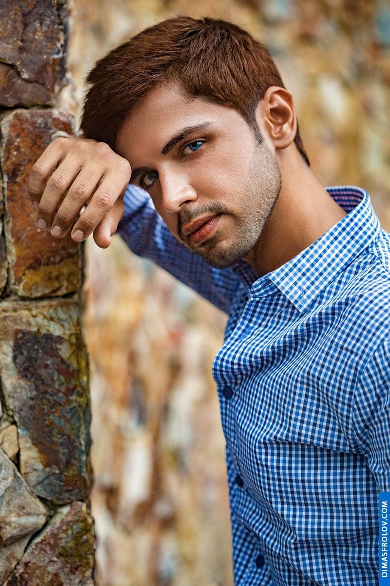 A rustic outdoor portrait: a man in a checkered shirt leans against a stone wall during a natural photo shoot